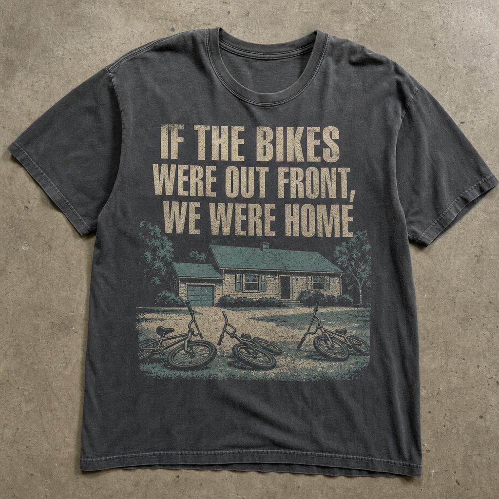 Washed charcoal t-shirt printed with faded cream and teal lettering above three children's bikes fallen on the lawn of a plain suburban house